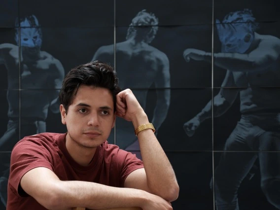 A man sits in front of a blue backdrop on which pictures of luche libre wrestlers are printed.